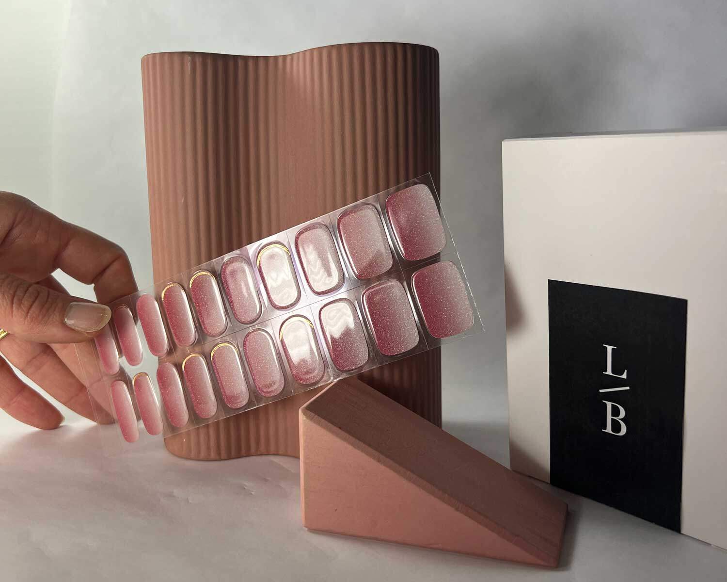 Golden sunset pink semi-cured gel nails in mockup to show product against wooden block and peach vase with la-bek uv lamp in the background hands holding nails