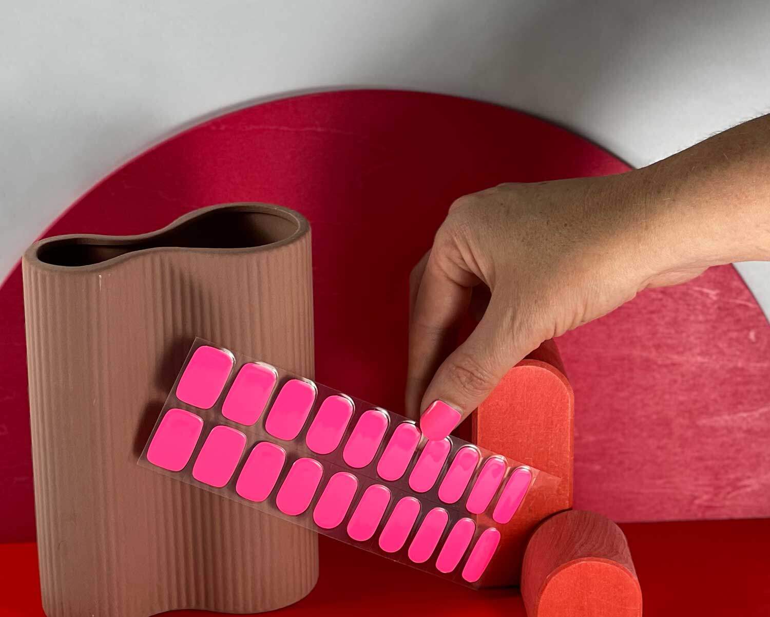 Barbie pink semi-cured gel nails in mockup to show product against red wooden blocks and peach vase with hand holding nail strip