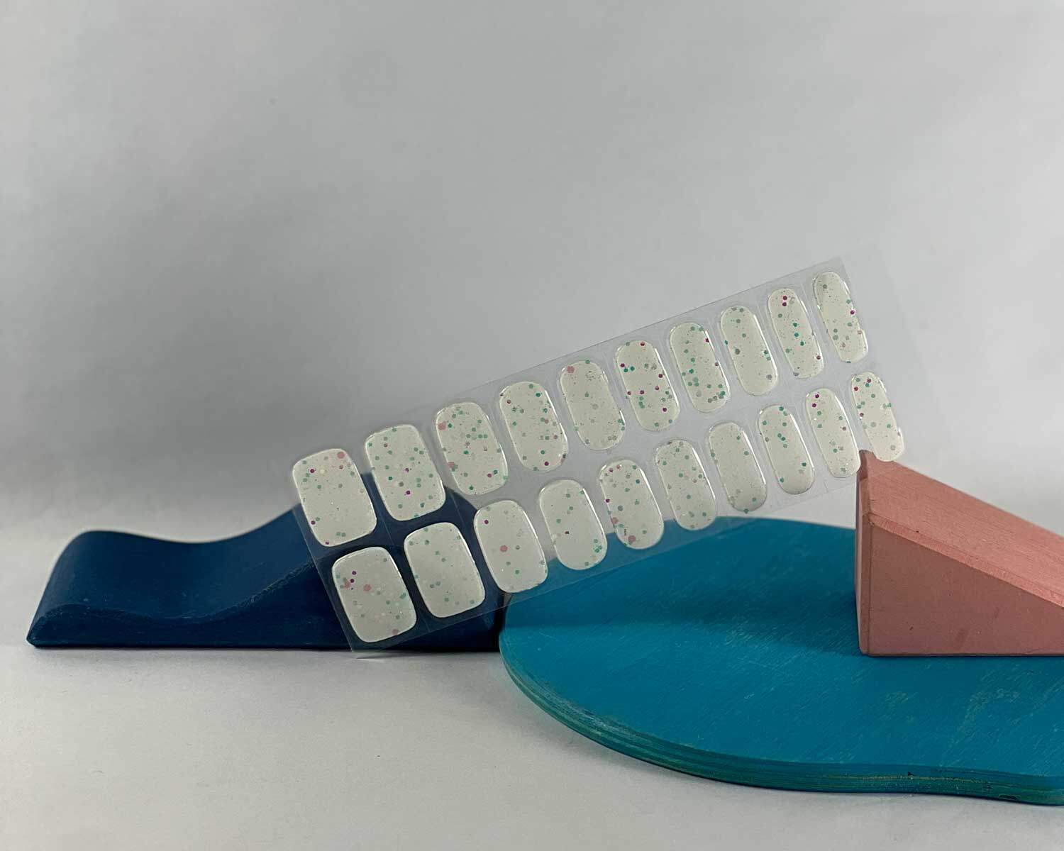 Sprinkle-Glow-Snow-semi-cured-gel-nail-mock Sprinkle snow Glow in the dark white semi-cured gel nails in mockup to show product against blue and pink wooden blocks