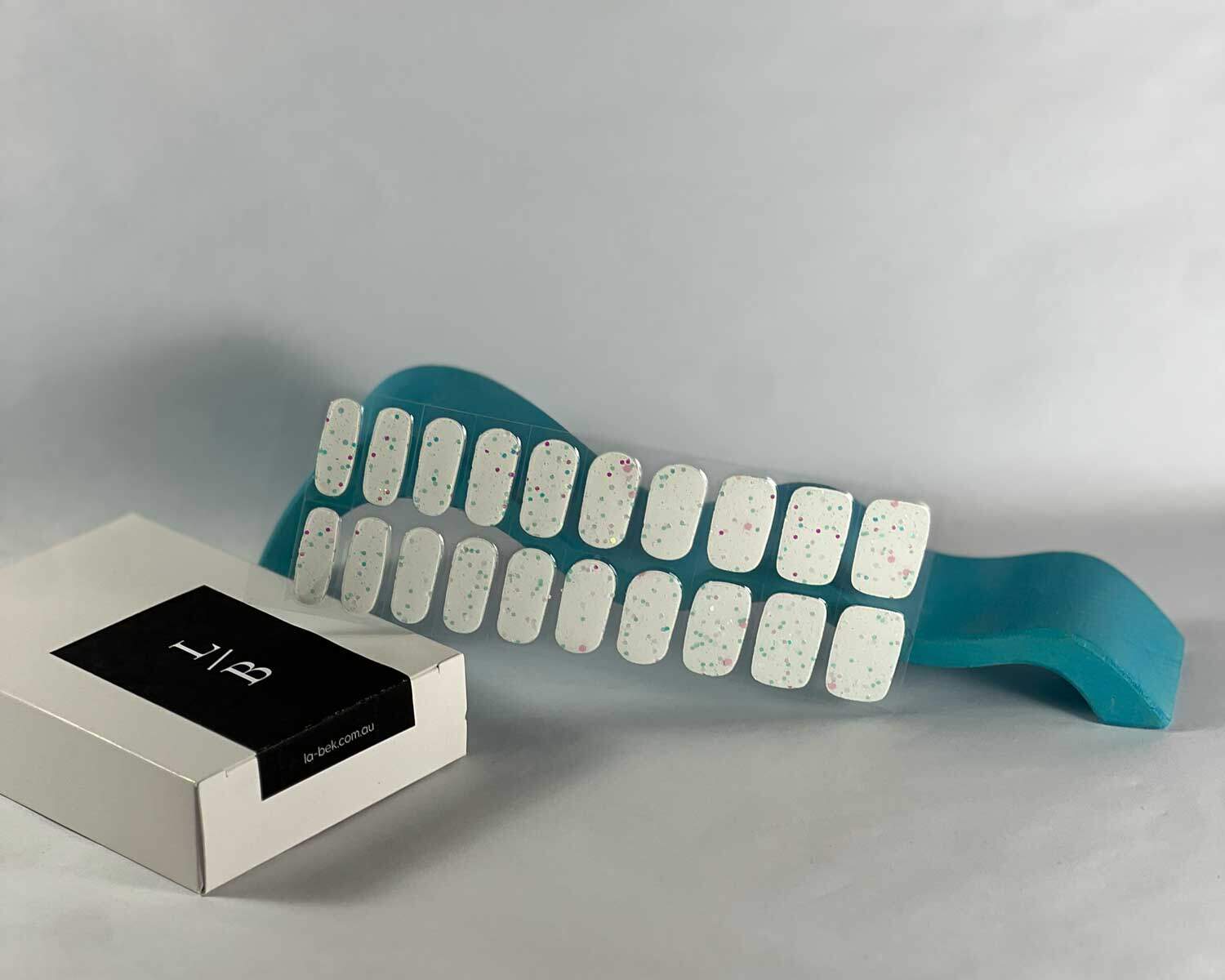 Sprinkle-Glow-Snow-semi-cured-gel-nail-curve Sprinkle snow Glow in the dark white semi-cured gel nails in mockup to show product against blue wave wooden block with la-bek uv lamp in the background