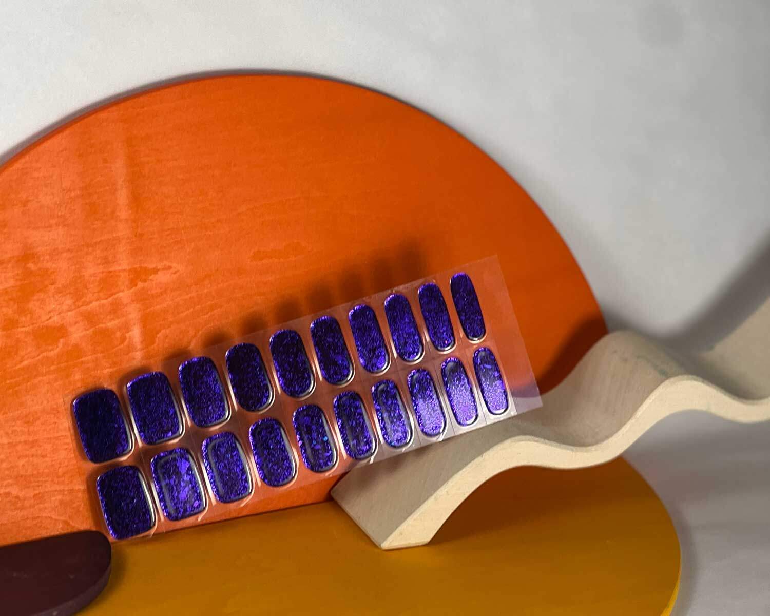 Violet Luster black and purple semi-cured gel nails in mockup to show product against white curved wooden block and yellow/orange blocks